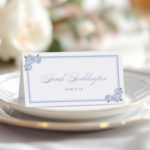 May include: A white place card with a blue border and floral accents. The text on the card reads "Sarah Reddington, Table 18."
