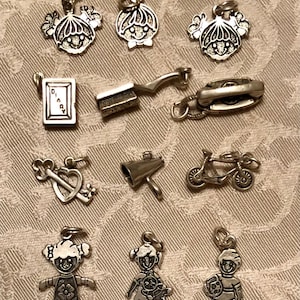 May include: A collection of silver-toned charms, including a girl with a flower dress, a boy with a football, a bicycle, a megaphone, a telephone, a diary, a hairbrush, and heart and key charms. The charms are on a textured background.