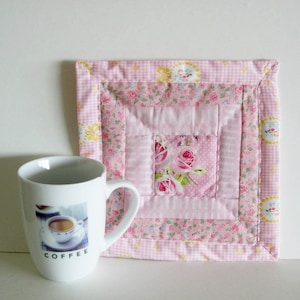 Könnte beinhalten: Eine weiße Kaffeetasse mit dem Aufdruck "COFFEE" neben einem rosa gesteppten Untersetzer. Der Untersetzer hat ein florales Rosendesign in der Mitte, umgeben von rosa und weiß gemusterten Stoffen. Die Tasse hat ein Bild von Kaffee.