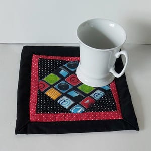 Könnte beinhalten: Ein weißer Keramikbecher steht auf einem quadratischen Stoffuntersetzer. Der Untersetzer hat einen rautenförmigen Mittelteil mit bunten App-Symbolen auf schwarzem Grund, umgeben von einem rot gepunkteten Rand und einem schwarzen Stoffrahmen.