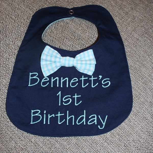 May include: A navy blue bib with a white and light blue bow tie and the text "Bennett's 1st Birthday" embroidered in light blue thread.