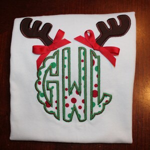 May include: White t-shirt with a green and red polka dot monogram surrounded by brown reindeer antlers and red bows.