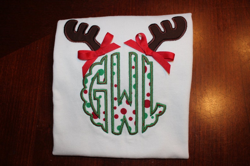 May include: White t-shirt with a green and red polka dot monogram surrounded by brown reindeer antlers and red bows.