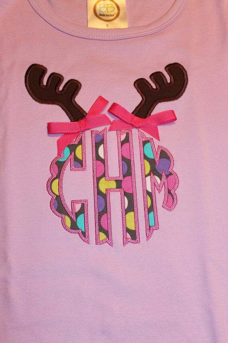 May include: A purple t-shirt with a brown reindeer applique. The reindeer has pink bows and a monogram with the letters "CHM" in a circle with a polka dot pattern.