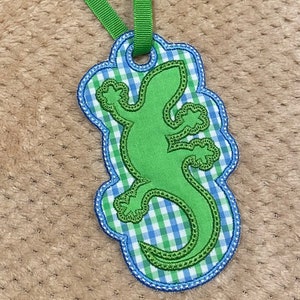 May include: A green lizard-shaped applique with a green ribbon. The applique is outlined in blue and features a gingham pattern in blue and green. The lizard is a lighter shade of green and has embroidered details.