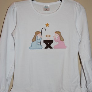 May include: White long-sleeved shirt with a ruffled hem and an embroidered nativity scene featuring two figures kneeling before a manger with a star above. The shirt has a tag that reads "GGG Girls" and "Size 8".