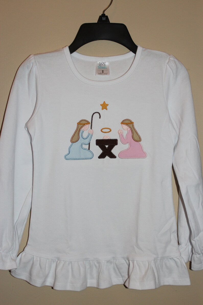 May include: White long-sleeved shirt with a ruffled hem and an embroidered nativity scene featuring two figures kneeling before a manger with a star above. The shirt has a tag that reads "GGG Girls" and "Size 8".