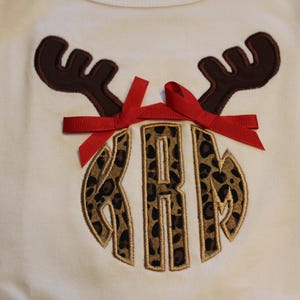 May include: A white shirt with a brown reindeer antler design and a red bow. The reindeer antlers are above a monogram with the letters "KRM" in a leopard print design.