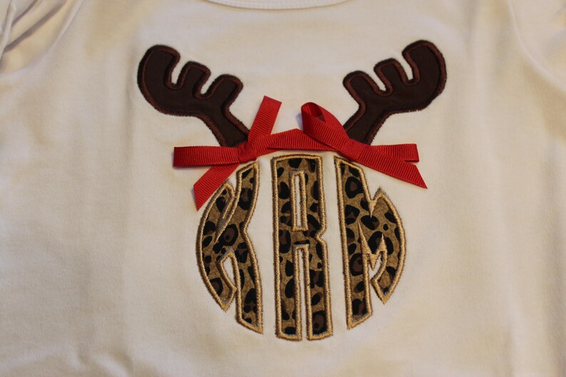 May include: A white shirt with a brown reindeer antler design and a red bow. The reindeer antlers are above a monogram with the letters "KRM" in a leopard print design.
