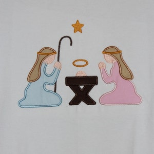May include: A white t-shirt with an embroidered nativity scene. The scene features a brown manger, a blue-robed figure kneeling on the left, a pink-robed figure kneeling on the right, and a gold star above. The text "CBB Blanks" and "8" are visible on the tag.