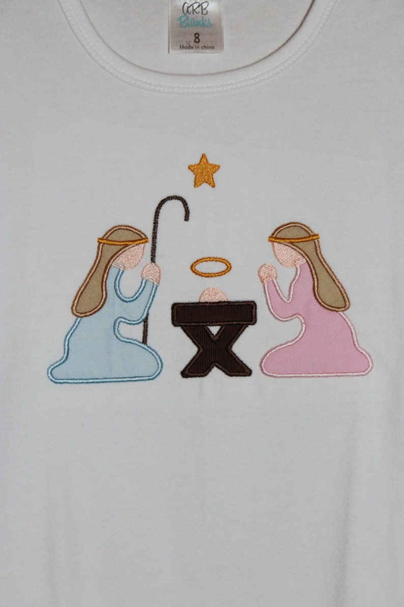 May include: A white t-shirt with an embroidered nativity scene. The scene features a brown manger, a blue-robed figure kneeling on the left, a pink-robed figure kneeling on the right, and a gold star above. The text "CBB Blanks" and "8" are visible on the tag.