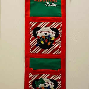 May include: A red, green, and white fabric wall hanging with a Mickey Mouse design. The hanging has three pockets and a wooden dowel at the top. The text "Very Merrytime Cruise" is embroidered on the top section.