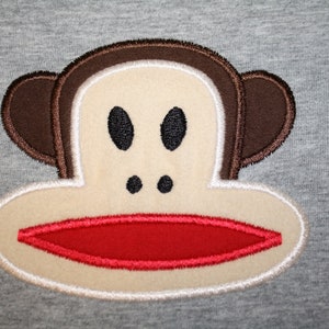 May include: Embroidered brown monkey face with a red mouth and white outline on a gray background.