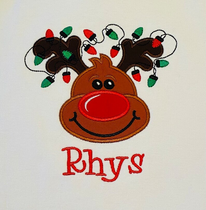 May include: A brown reindeer with a red nose and green and red lights around its antlers. The name Rhys is embroidered below the reindeer.