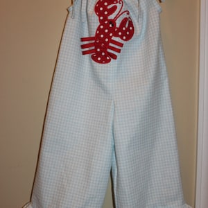 May include: A light blue and white gingham jumpsuit with a red and white lobster applique. The jumpsuit has a ruffled hem and straps.