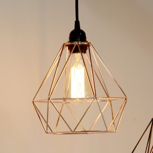 Puede incluir: Una lámpara colgante geométrica con una estructura metálica de color cobre. La bombilla es visible dentro del marco. La lámpara cuelga de un cable negro y un soporte de techo negro. El diseño es moderno e industrial.