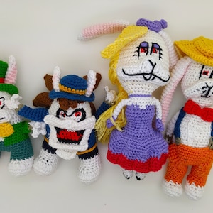 Peut inclure: Quatre poupées lapins crochetées à la main, vêtues de différentes tenues. L'une porte un chapeau haut de forme et une veste verts, une autre une tenue bleue et marron, une troisième une robe violette et la dernière une tenue orange. Chacune a des yeux rouges.