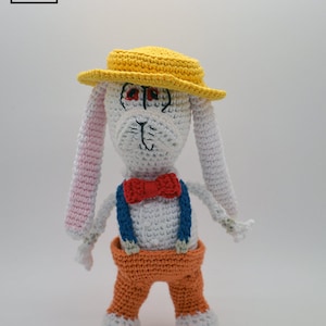 Könnte beinhalten: Ein weißer Häkelhase mit einem gelben Hut, einer orangefarbenen Hose und einer roten Schleife. Der Hase hat rosa Ohren und steht auf einem weißen Hintergrund.