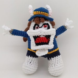 Peut inclure: Poupée au crochet faite à la main d'un personnage de dessin animé avec un visage blanc, des yeux rouges et une bouche grande ouverte. La poupée a un chapeau bleu, des cheveux bruns et des cornes blanches. Elle porte une tenue bleue et blanche avec des gants blancs.
