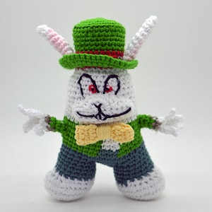 Puede incluir: Un conejo blanco de crochet que lleva un sombrero verde con una banda roja, un chaleco verde, una pajarita amarilla y pantalones azules. El conejo tiene ojos rojos y una sonrisa traviesa.