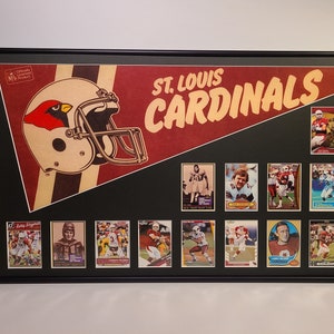 May include: Framed St. Louis Cardinals memorabilia. The artwork features a vintage-style pennant with a football helmet and the team name. Below the pennant are multiple football trading cards, all encased in a black frame.