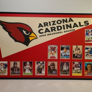 May include: Framed display featuring Arizona Cardinals memorabilia. The design includes the team logo, text reading "ARIZONA CARDINALS 2006 INAUGURAL SEASON", and a collection of football trading cards. The frame is black, and the background is red.
