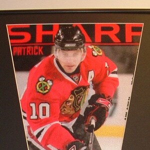 May include: A framed black and white photo of a hockey player in a red and white jersey with the name "Patrick Sharp" on the jersey. The photo is surrounded by smaller photos of the same player in different poses. The photo is framed in a black frame with a white mat.