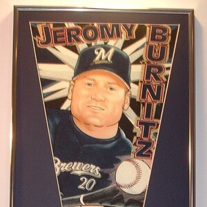 May include: A framed pennant with a blue background and a white triangle. The triangle features a baseball player wearing a blue jersey with the word "Brewers" and the number "20" on it. The pennant also features the player's name "JEROMY BURTIZ" in white letters. The pennant is surrounded by four baseball cards featuring the same player.