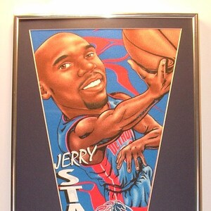 May include: A framed pennant featuring a cartoon illustration of Jerry Stackhouse, a basketball player for the Detroit Pistons. The pennant is blue and red with the words "Jerry Stackhouse" and "Pistons" on it. The frame is silver and has a blue background. There are also four trading cards of Jerry Stackhouse in the frame.
