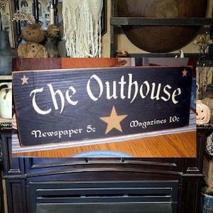 May include: A black wooden sign with white lettering that reads "The Outhouse" with a gold star and the text "Newspaper 5c" and "Magazines 10c".