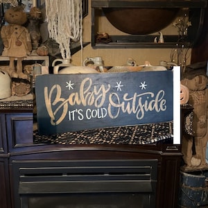 May include: A black wooden sign with white lettering that reads "Baby It's Cold Outside". The sign is decorated with white snowflakes.