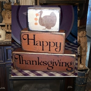Puede incluir: Un letrero de madera de tres niveles con las palabras "Happy Thanksgiving" y una silueta de un pavo con bayas naranjas. Los bloques están pintados en tonos marrones y naranjas con letras negras.