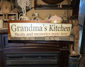 May include: A rustic wooden sign with the text "Grandma's Kitchen Meals and memories made here" in black lettering on a white background. The sign is hanging above a fireplace.