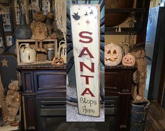 May include: A white wooden sign with red lettering that reads "SANTA Stops Here". The sign has a blue and green background with a silhouette of Santa Claus and reindeer flying through the air.