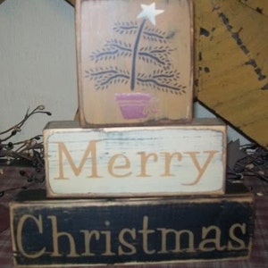 May include: Three wooden blocks stacked on top of each other. The top block is beige with a black silhouette of a Christmas tree with a white star on top. The middle block is white with the word "Merry" in black. The bottom block is black with the word "Christmas" in white.