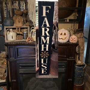 May include: A black wooden sign with white lettering that reads "FARMHOUSE" with a stylized sunburst design in the center. The sign is leaning against a fireplace with a decorative metal container and a rustic wooden bucket.