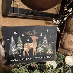 Puede incluir: Un letrero rectangular de madera negra con una escena invernal. Presenta un ciervo con un diseño estampado, árboles estilizados y copos de nieve. El texto "walking in a winter wonderland" está en la parte inferior. El letrero está decorado con colores blanco y marrón.