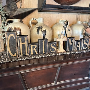 Puede incluir: Bloques de madera negros que deletrean "NAVIDAD" con una cruz dorada en el centro. Los bloques están sobre una superficie de madera marrón oscuro frente a una colección de jarras de gres antiguas.