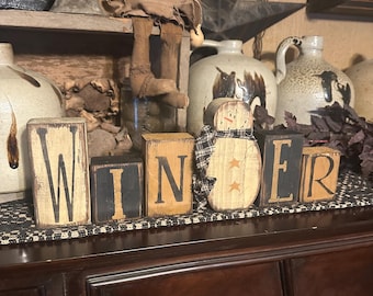 May include: Wooden blocks spelling "WINTER" with a snowman figure. The letters are black on a cream background. The snowman has a plaid scarf and star accents. The blocks sit on a dark wooden surface.