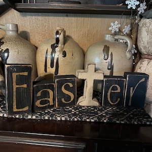 May include: Black wooden blocks spelling out the word "Easter" with a white wooden cross in the centre. The blocks are sitting on a brown wooden surface.