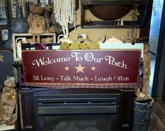 May include: A red and gold wooden sign with the text "Welcome to Our Porch Sit Long - Talk Much - Laugh Often". The sign has a plaid border and three gold stars.