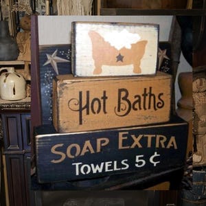 May include: Three wooden signs stacked on top of each other. The top sign is white with a black silhouette of a bathtub with bubbles. The middle sign is brown with black text that says "Hot Baths". The bottom sign is black with white text that says "Soap Extra Towels 5¢".
