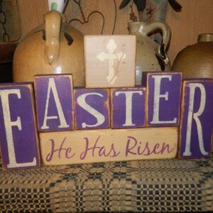 May include: Wooden block letters spelling "Easter" in white letters on a purple background. A smaller block below reads "He Has Risen" in white letters on a beige background. The blocks are arranged on a brown and white patterned surface.