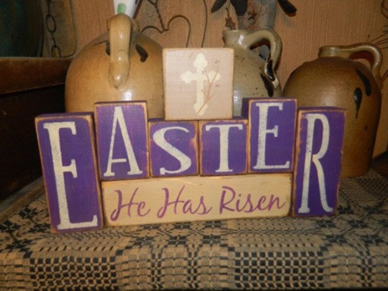 May include: Wooden block letters spelling "Easter" in white letters on a purple background. A smaller block below reads "He Has Risen" in white letters on a beige background. The blocks are arranged on a brown and white patterned surface.