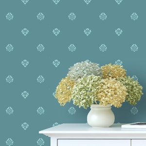 May include: A white vase holding a bouquet of dried flowers in shades of gold, green, and silver, set against a teal wall with a repeating floral pattern. The vase sits on a white table.