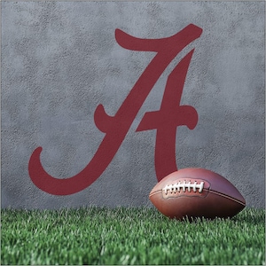 May include: A brown leather football rests on green grass in front of a gray wall with a large crimson letter "A". The football has white laces. The image evokes a sense of sports and team spirit.