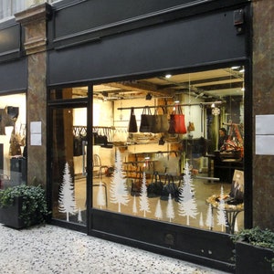 May include: A storefront window displays a collection of handbags in various styles and colors. The window is decorated with white tree silhouettes. The shop's interior is visible, with bags hanging from racks and shelves.