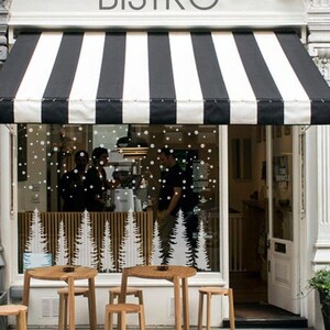 May include: A storefront window with a black and white striped awning. The window is decorated with a winter scene featuring white snow and pine trees. There are four tables and chairs outside the store.