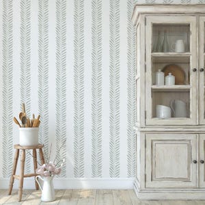 May include: A white wall with vertical gray leaf pattern wallpaper. A wooden stool holds a white pot with wooden utensils. A white pitcher with pink flowers sits on the floor. A white cabinet with glass doors is on the right.