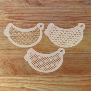 May include: Three light-colored, curved stencils with a handle-like loop on each end. The stencils feature different patterns, including a fish scale design and a geometric pattern. The stencils are arranged on a wooden surface.
